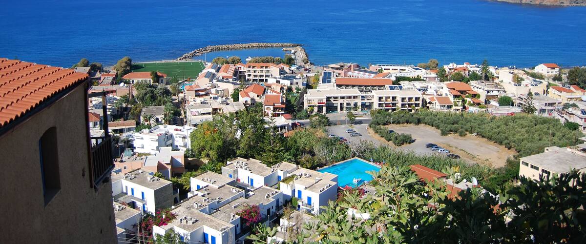 A coastal town with white buildings, a blue pool, and a rocky island in the distance.