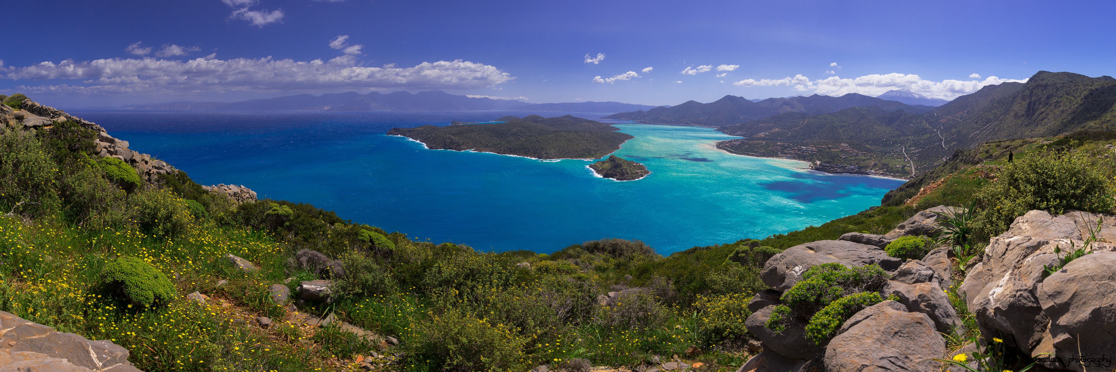 A coastal landscape with a clear blue sea, green hills, and rocky terrain.