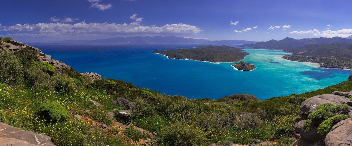 A coastal landscape with a clear blue sea, green hills, and rocky terrain.