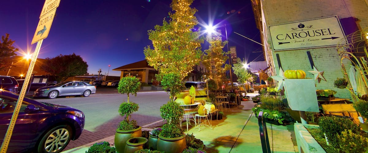 A nighttime street scene with a decorated Christmas tree, parked cars, and a building with a sign reading "CAROUSEL French Cuisine."