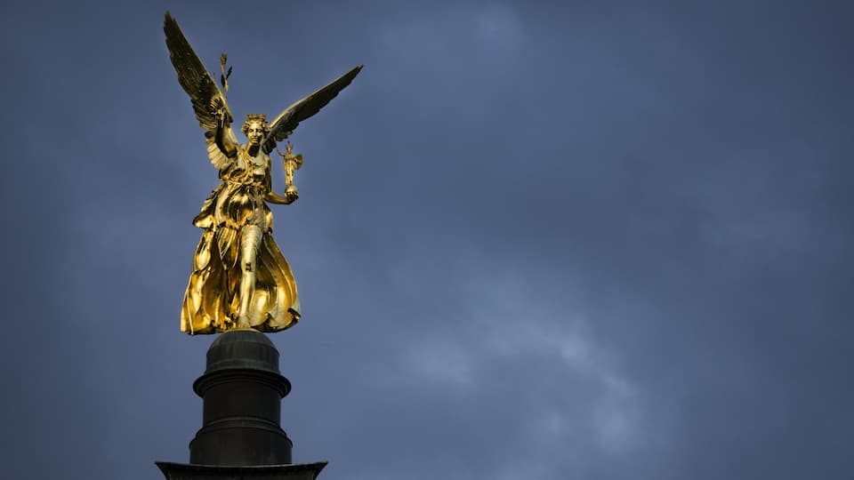 A golden angel statue atop a dark pedestal against a cloudy sky.