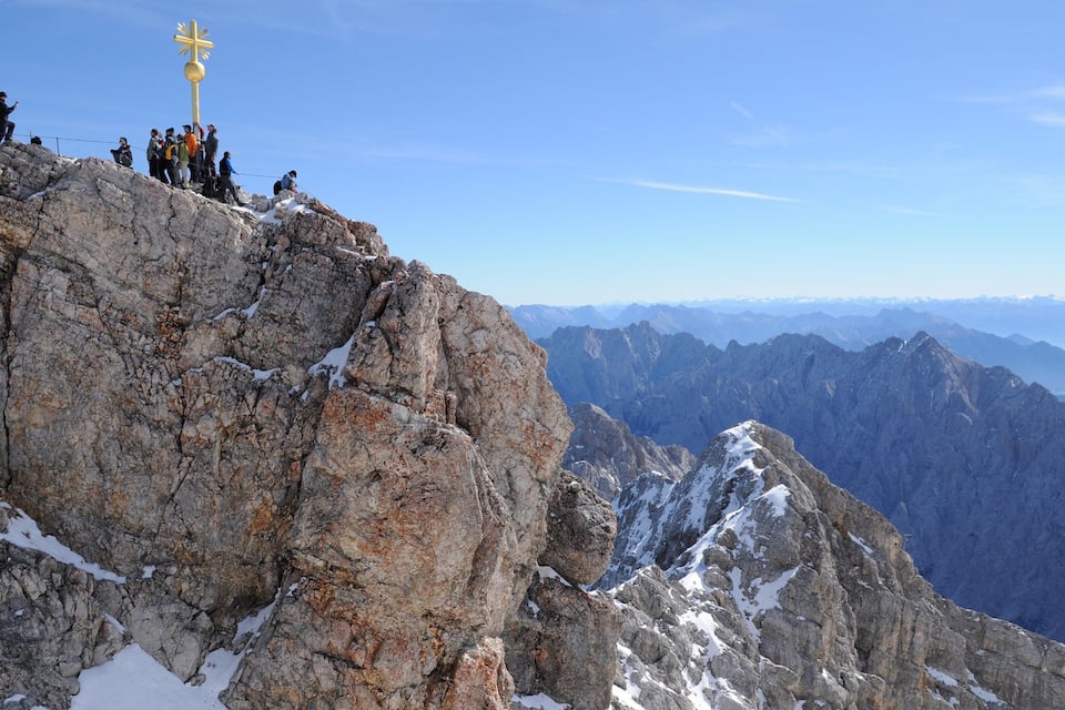 A group of hikers on a rocky mountain peak with a golden cross, surrounded by snow-covered slopes and distant mountain ranges.
