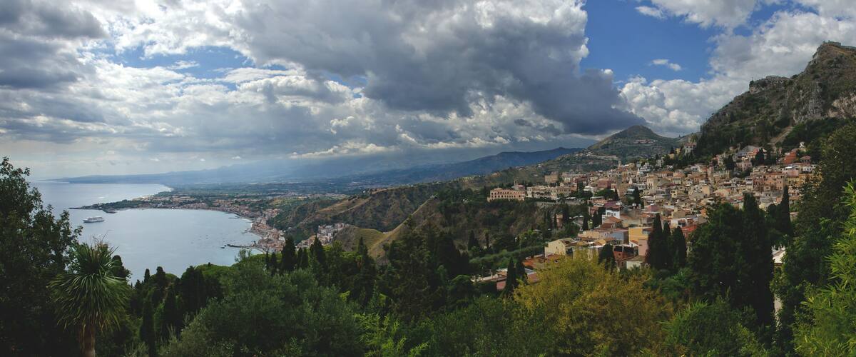 A coastal town nestled in a mountainous area with a prominent castle on a hill and a large body of water in the foreground.