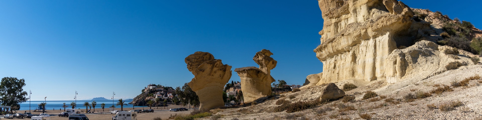 Paisagem costeira com formações rochosas imponentes e uma área de praia com veículos estacionados.