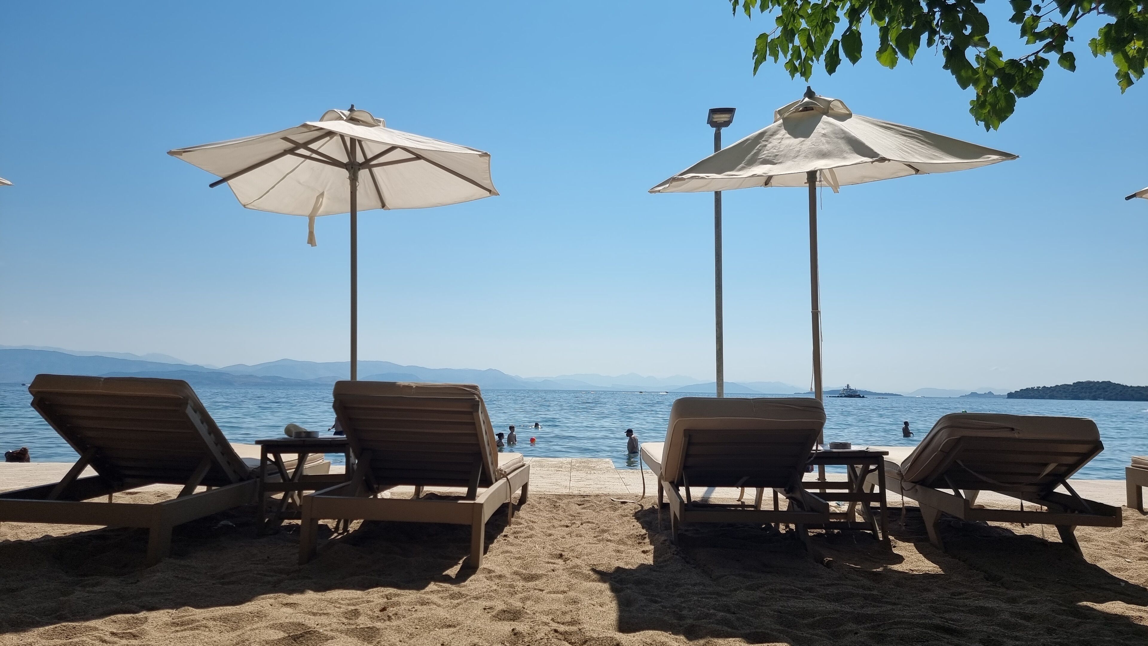 Una playa con tumbonas y sombrillas, un mar tranquilo y montañas a lo lejos.
