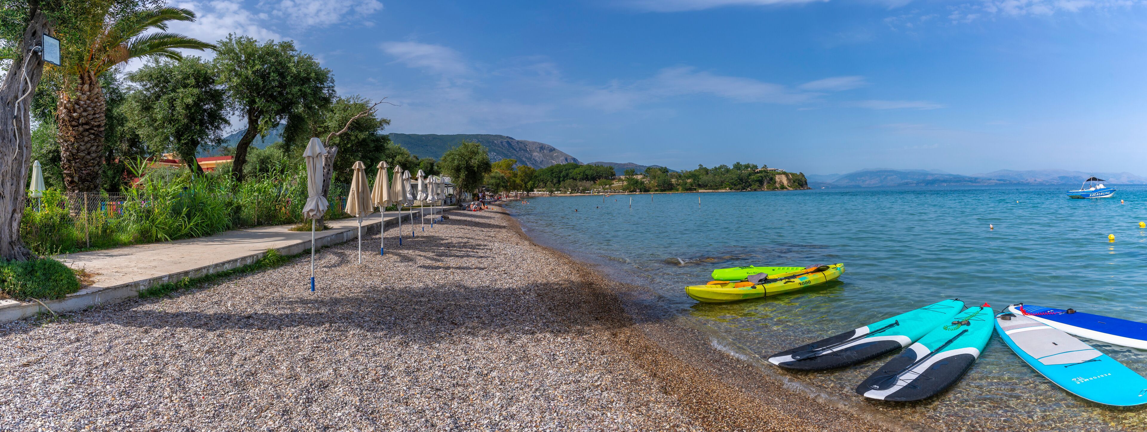 A pebble beach with kayaks and a boat on calm water, palm trees, and a distant island.