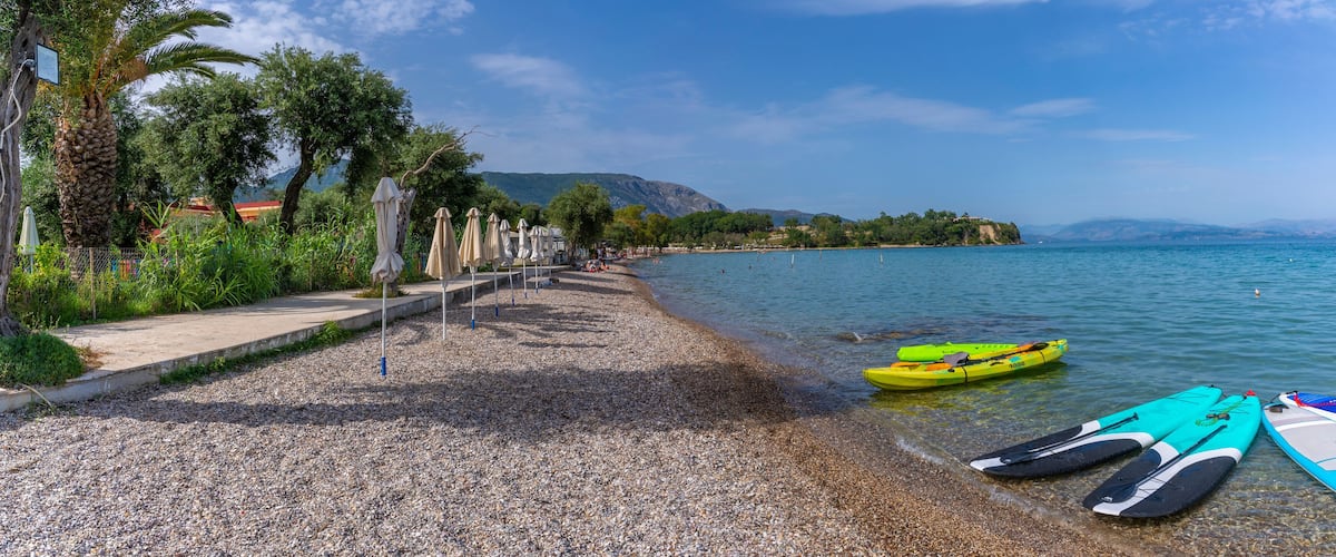 A pebble beach with kayaks and a boat on calm water, palm trees, and a distant island.