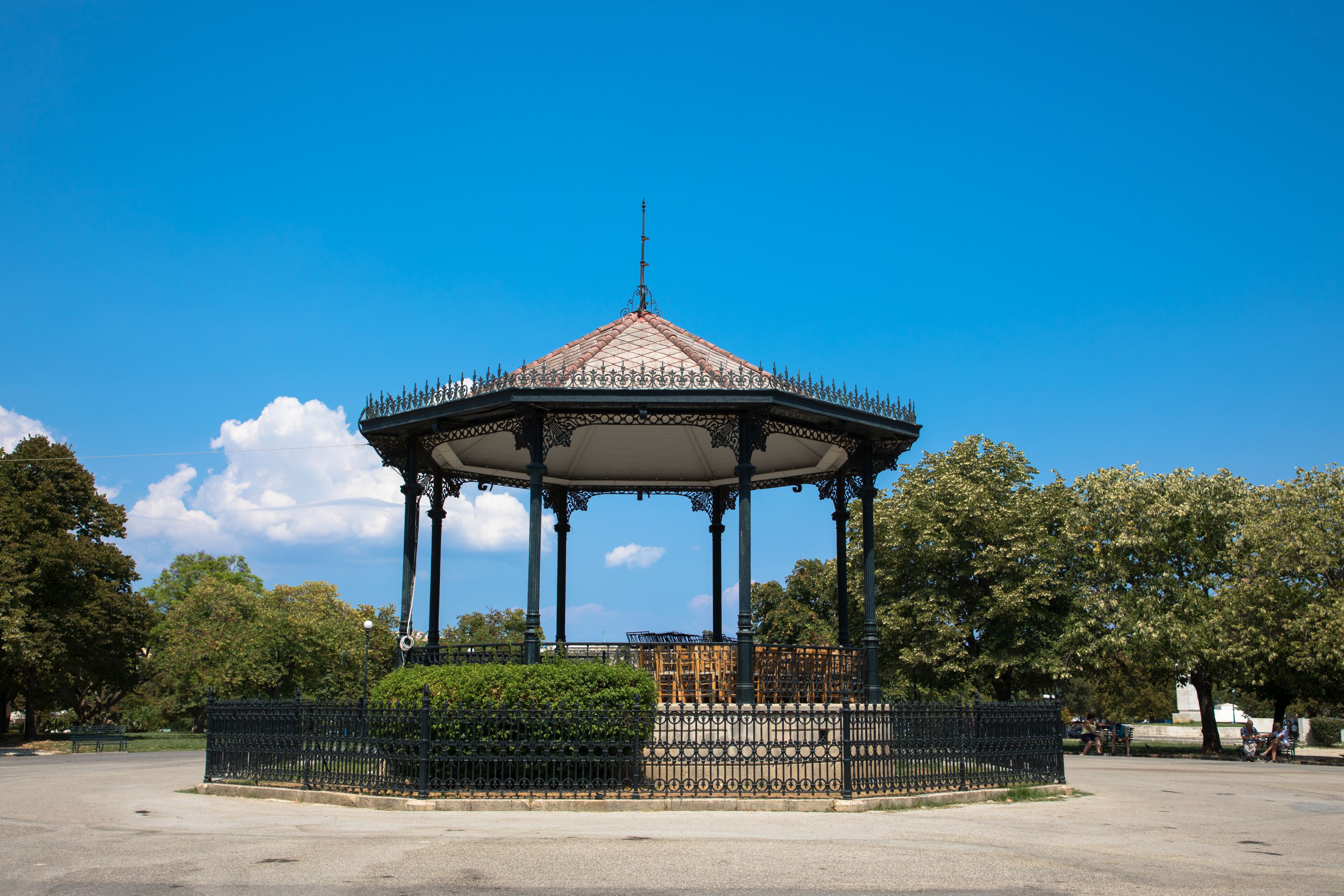 Un pabellón con detalles metálicos decorativos y techo de tejas, rodeado de una valla y árboles en un parque.