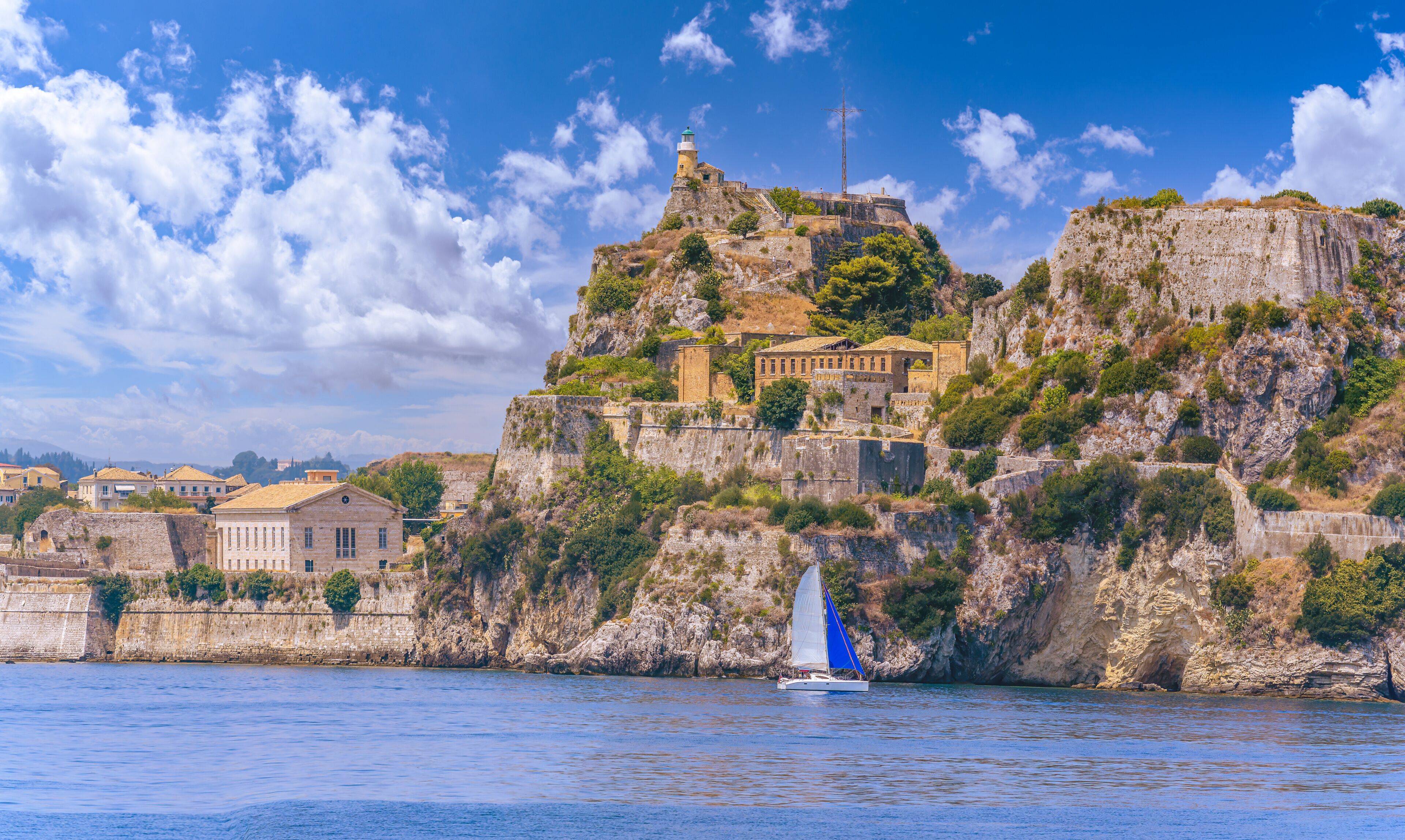 A coastal town with historic buildings perched on a cliff, a lighthouse, and a sailboat on the water.