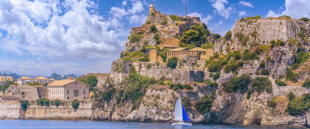 A coastal town with historic buildings perched on a cliff, a lighthouse, and a sailboat on the water.