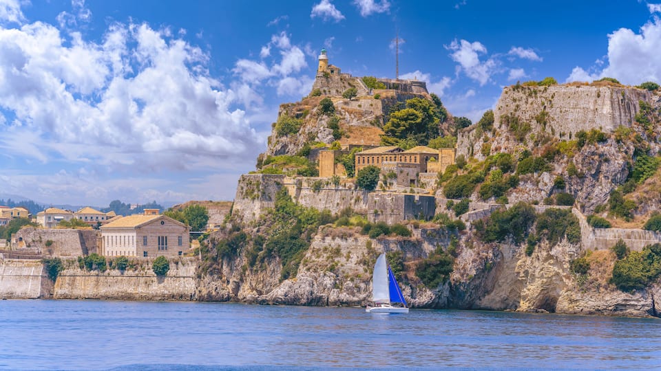 A coastal town with historic buildings perched on a cliff, a lighthouse, and a sailboat on the water.