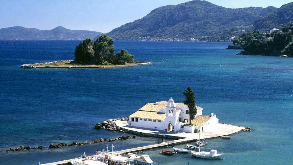 A coastal town with a white church, boats docked at the pier, and a small island with trees.