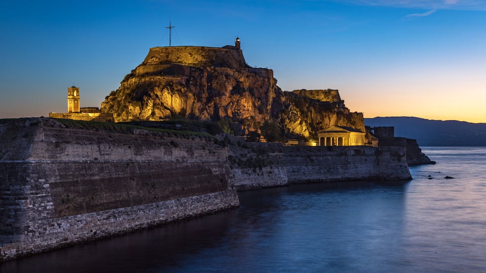 A fortified structure with a lighthouse on a cliff overlooking calm water.