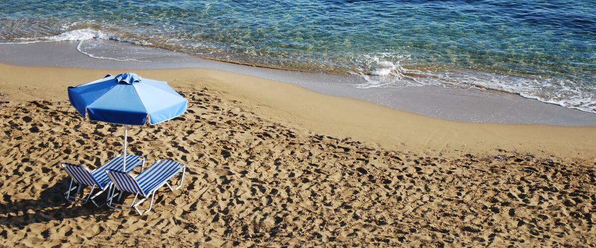 A sandy beach with clear blue water, a blue umbrella, and two striped lounge chairs.