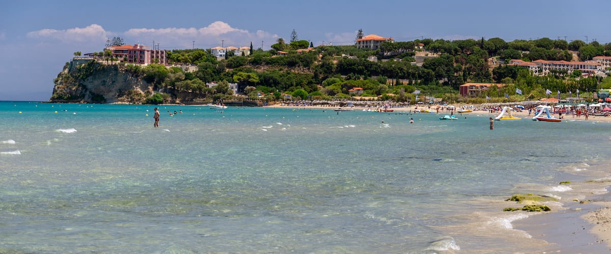 A coastal beach with clear turquoise waters, a sandy shore, and buildings on a hillside in the background.
