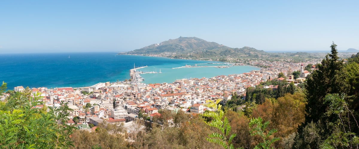 A coastal town with a mountain in the background, a clear blue sea, and dense greenery.
