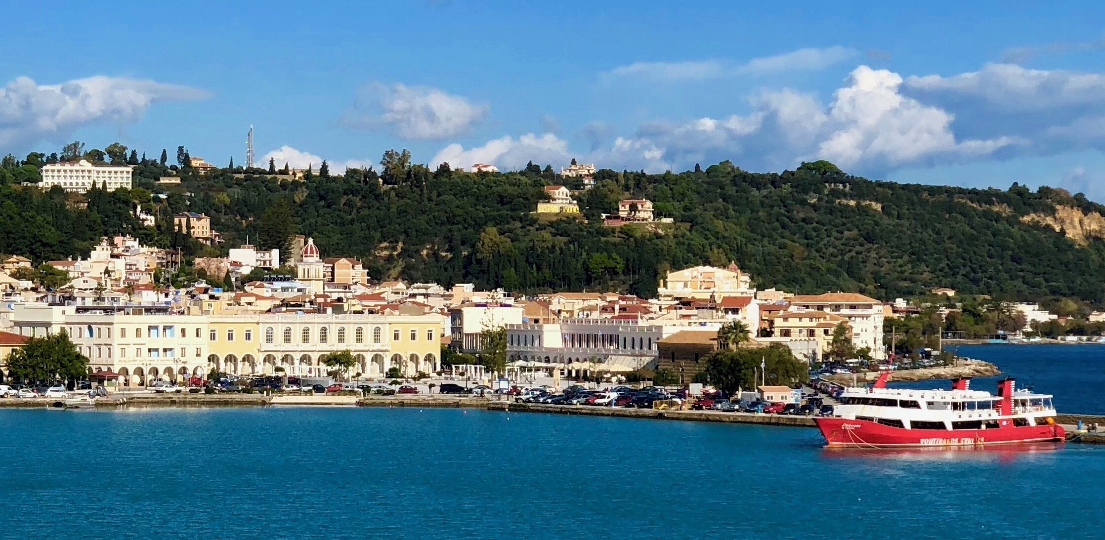 A coastal town with a prominent red ferry, historic buildings, and a hilly backdrop.