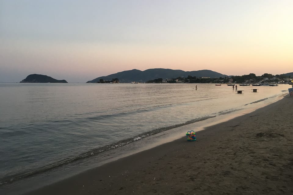 Uma praia com águas calmas, uma ilha ao longe e algumas pessoas distantes.