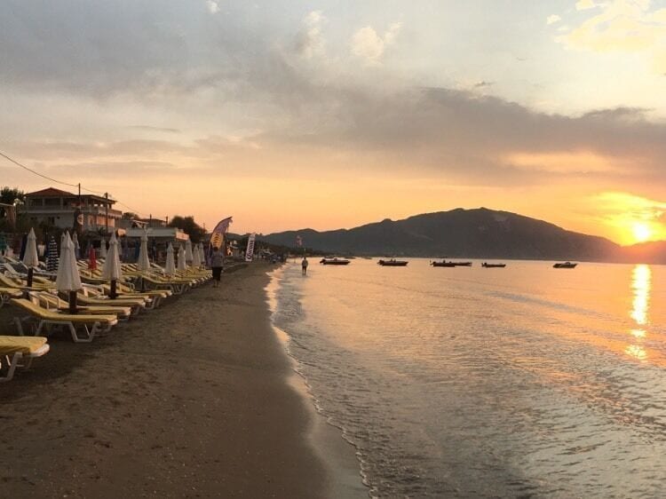 A beach with sun loungers and umbrellas, boats on the water, and a mountain in the background.