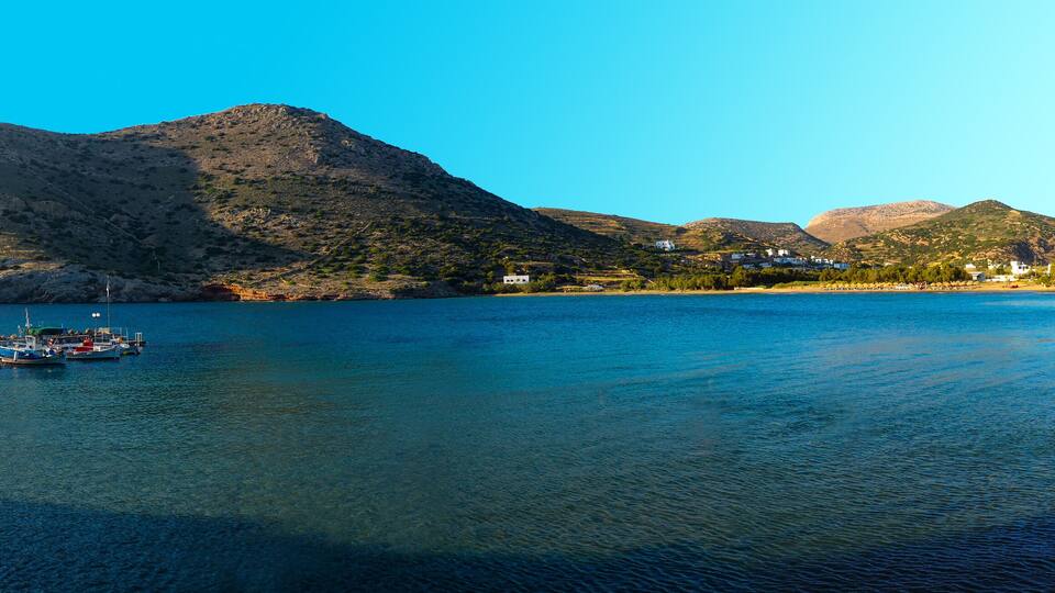 A calm lake with boats docked on the left, surrounded by rocky hills and white buildings.