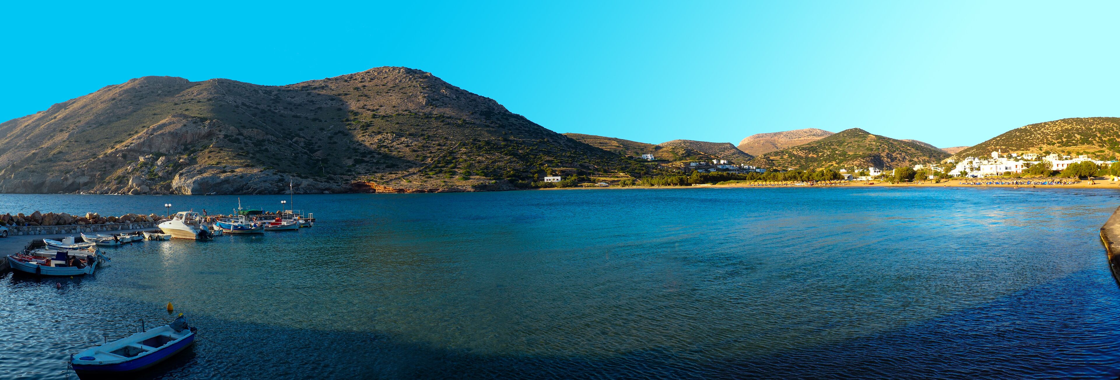 A calm lake with boats docked on the left, surrounded by rocky hills and white buildings.