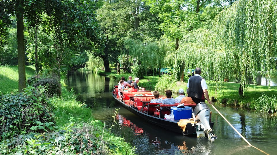 A narrow waterway lined with lush greenery and willow trees, with a boat carrying passengers.