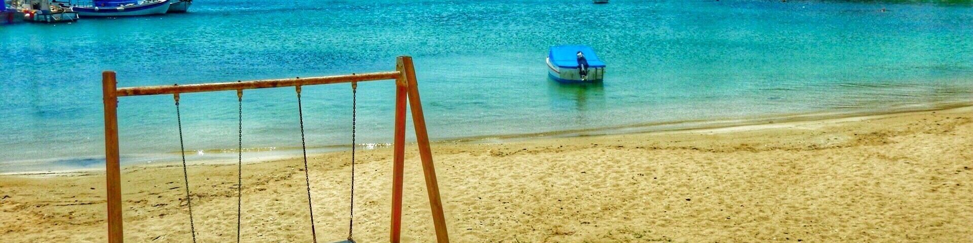 Uma praia com baloiço, águas turquesa límpidas, barcos e edifícios ao fundo.