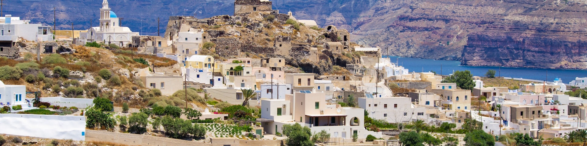 A coastal village with white buildings, a church with a blue dome, and a rocky cliff by the sea.