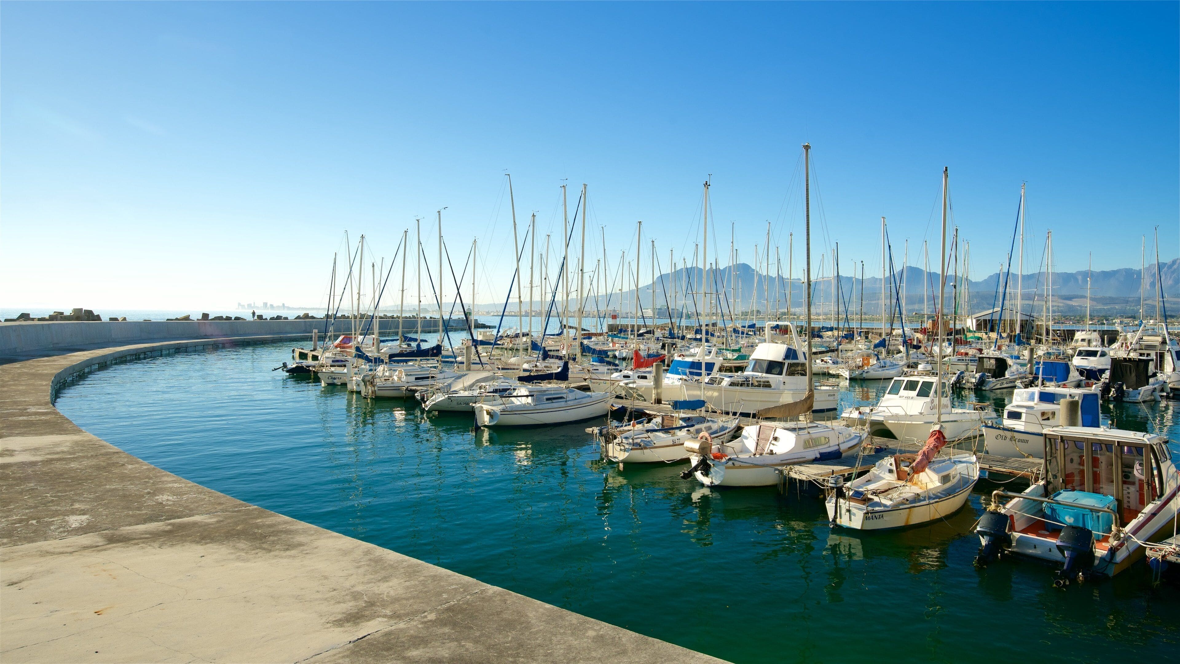 A marina with numerous sailboats docked along a curved concrete quay, with mountains in the background.
