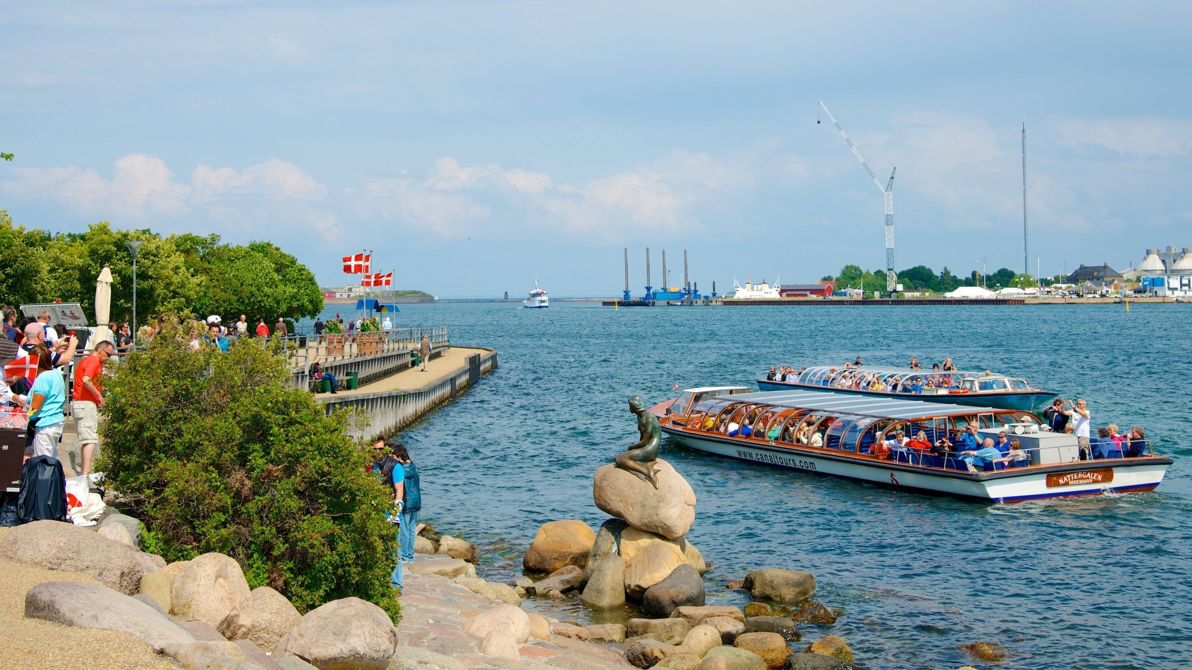 A boat tour with passengers, a pier with people, and industrial structures in the background.