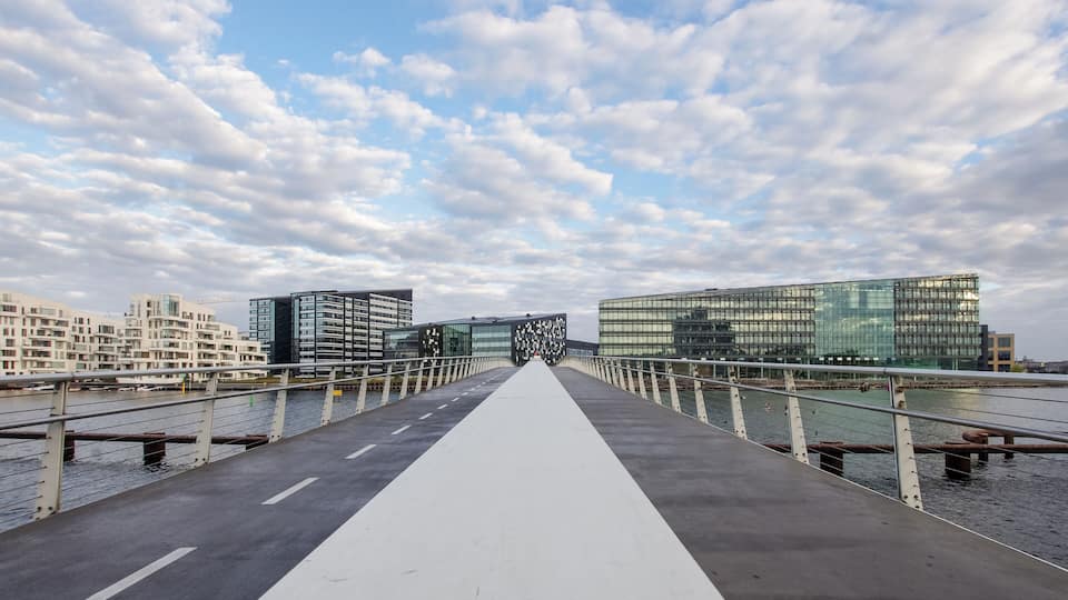 A modern bridge with a white center path, flanked by water on both sides and buildings in the background.