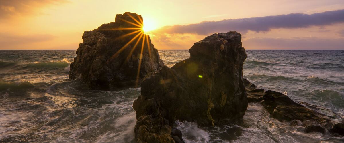 A sunset over rocky coastal waters with sunlight shining through.