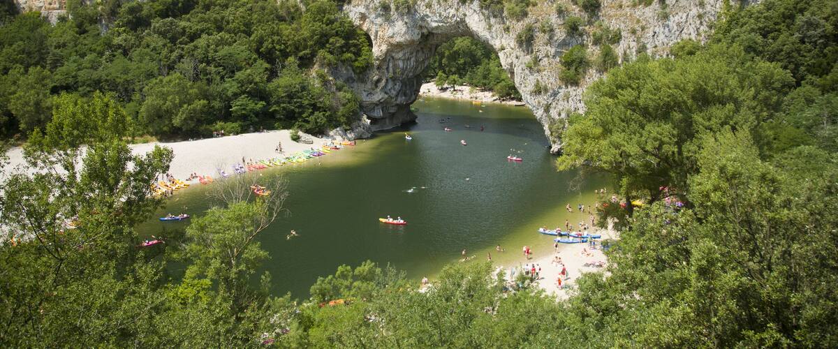 A natural arch formation over a river surrounded by dense forest and a sandy beach.