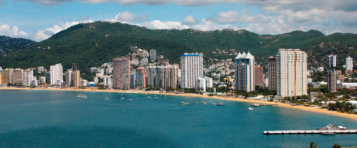 A coastal city with a beach, boats, and a mountainous backdrop.