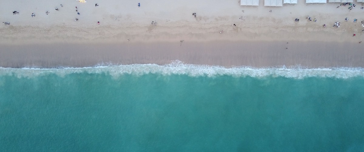 An aerial view of a beach with clear turquoise water, a sandy shore, and people scattered along the beach.
