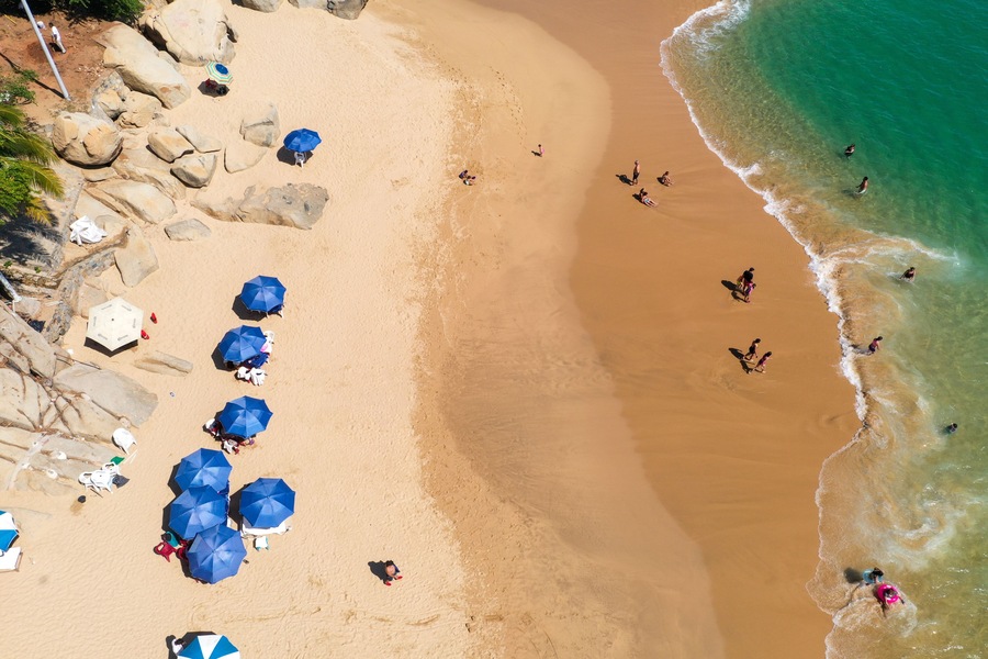 Una playa arenosa con rocas grandes, varias sombrillas azules y gente nadando en el agua.