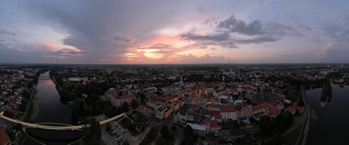 A panoramic view of a town at dusk with a river running through it, surrounded by buildings and a bridge.
