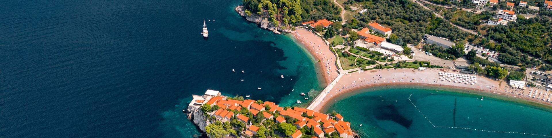 An aerial view of a coastal town with a prominent rocky peninsula, clear blue waters, and a beach area with buildings and boats.