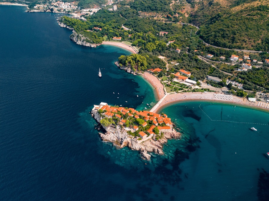 Vista aérea de un pueblo costero con un pronunciado promontorio rocoso, aguas azules cristalinas y una zona de playa con edificios y barcos.