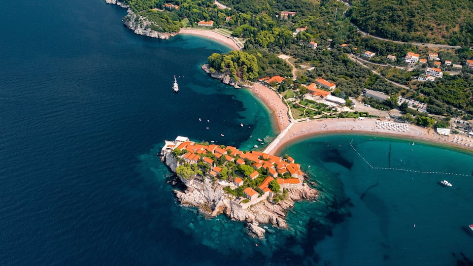 An aerial view of a coastal town with a prominent rocky peninsula, clear blue waters, and a beach area with buildings and boats.