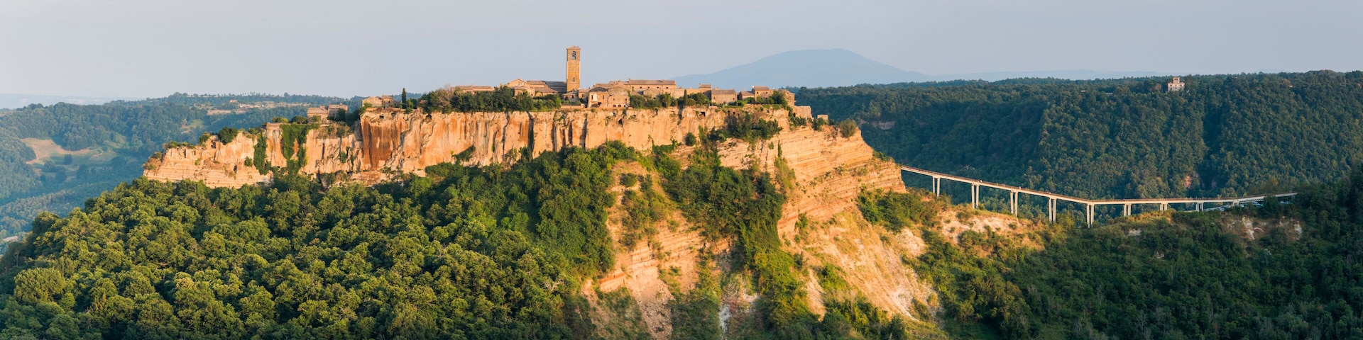 A mountainous landscape with a historic fortress atop a cliff, a bridge spanning a valley, and dense forested hills in the background.