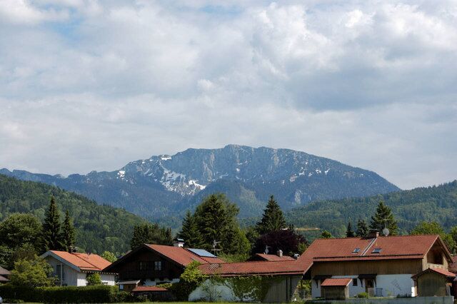 A mountain range with snow-capped peaks, houses with red roofs, and lush green trees in the foreground.