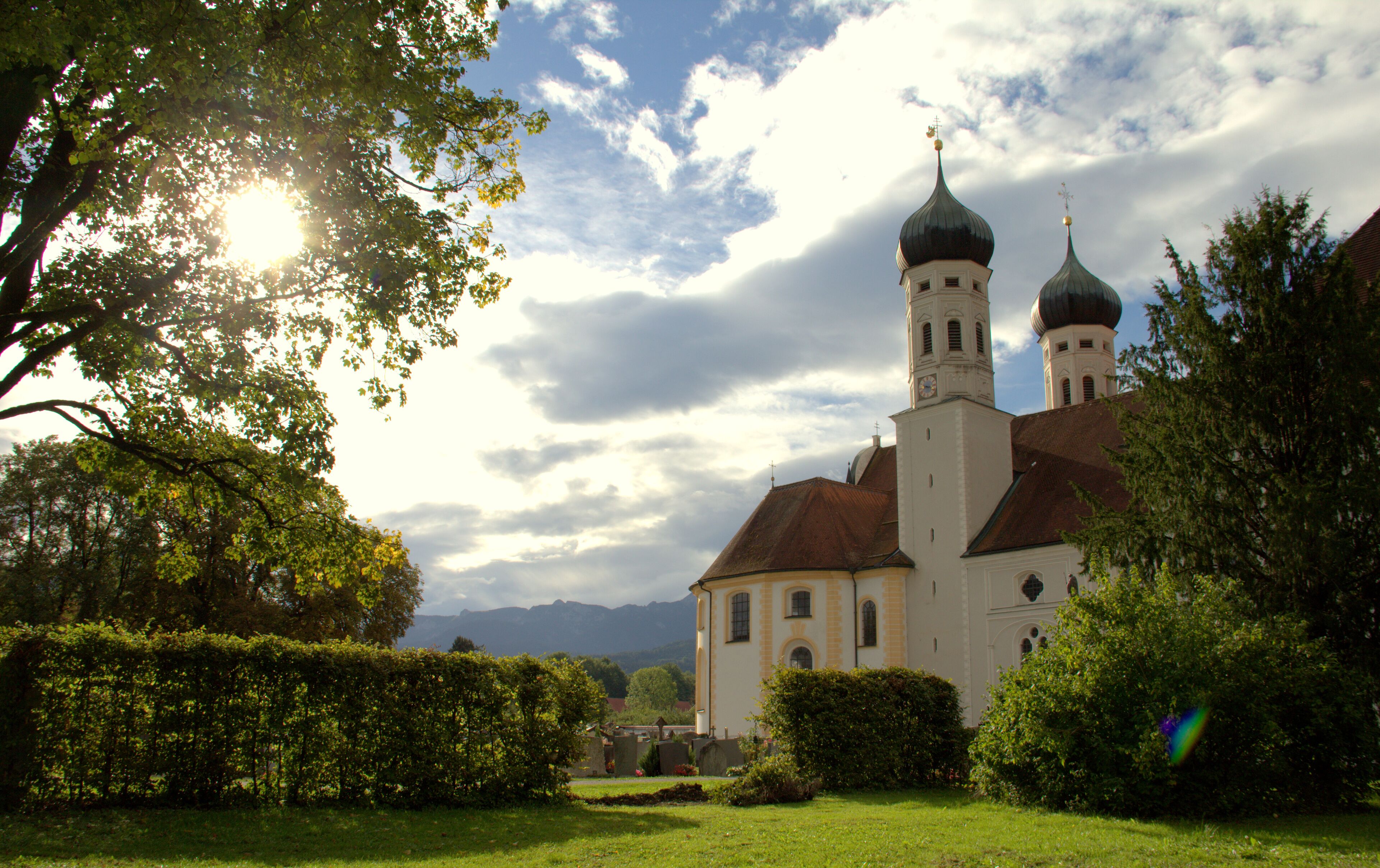 A church with two domes and a bell tower, surrounded by greenery and mountains in the background.