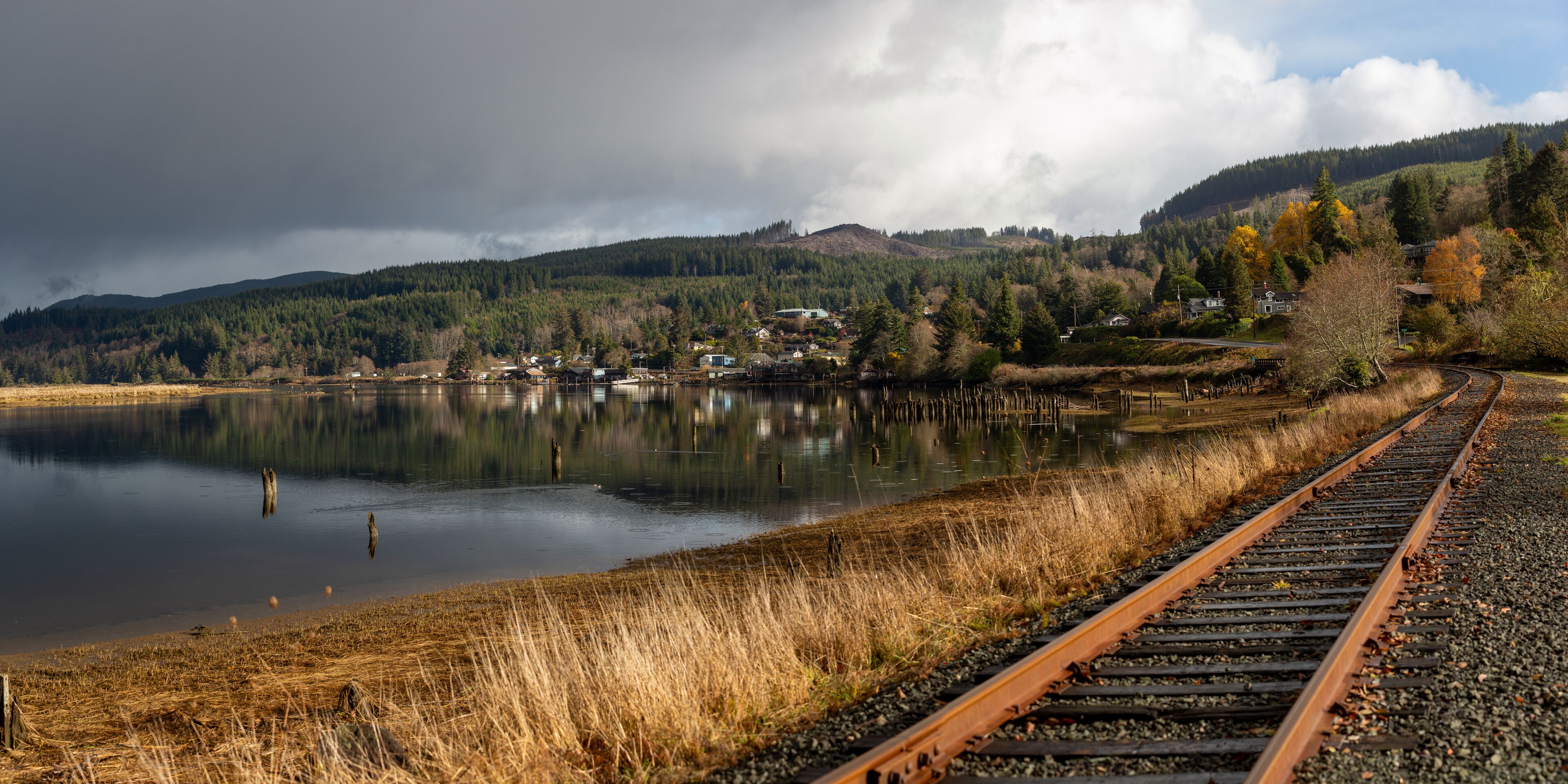 A lake with a railway track, surrounded by forested hills and houses.