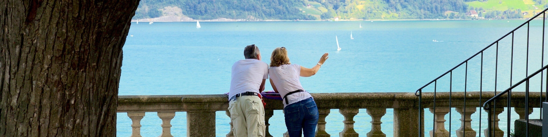 Un couple se tient sur un promontoire surplombant un lac où naviguent des voiliers, le tout étant entouré de collines.