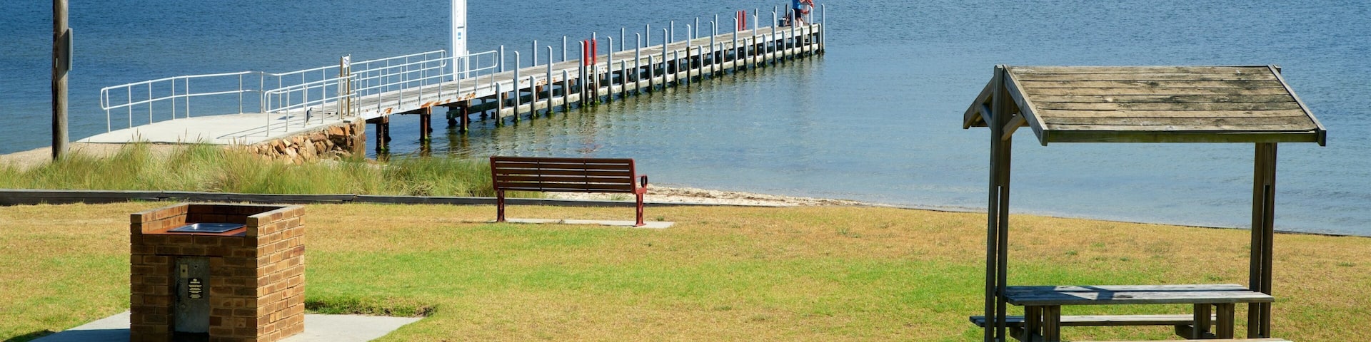 A pier extending into a calm body of water, a wooden bench, and a picnic shelter on a grassy area.