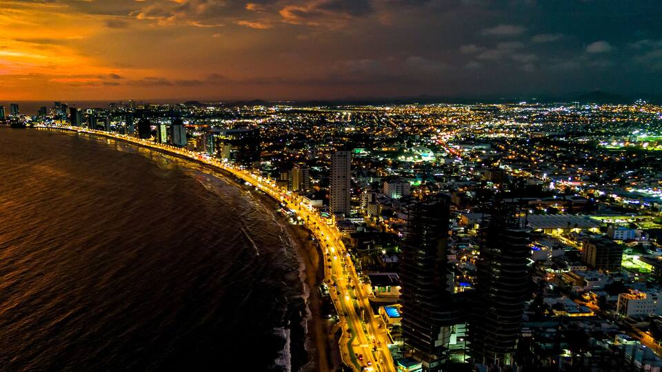 A coastal cityscape at sunset with a brightly lit beach and dense urban area.