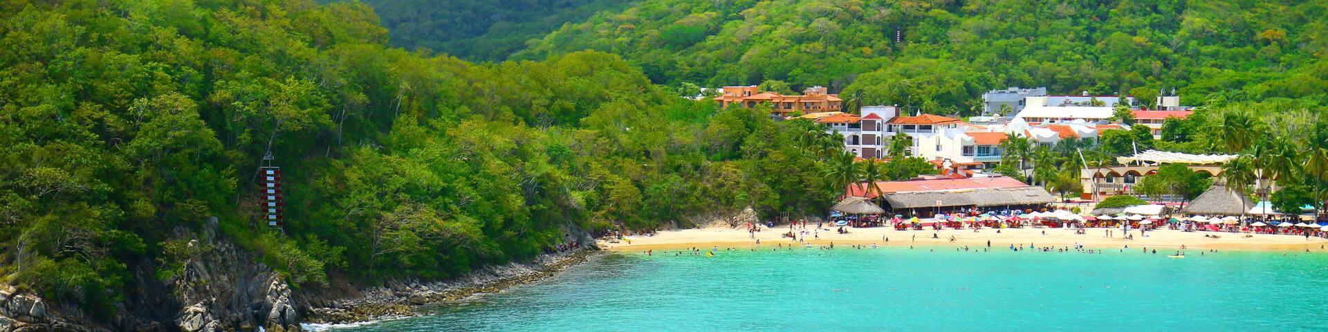 A turquoise sea with a wake, a beach with people, and buildings on a hillside.