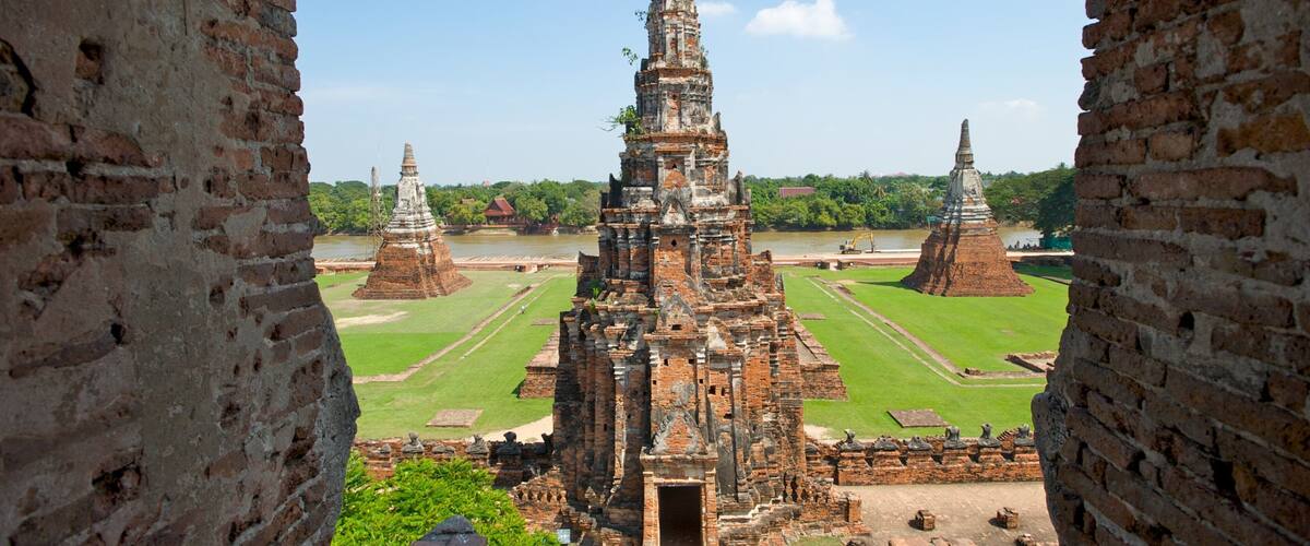 An ancient temple complex with multiple structures, including a central towering temple and smaller surrounding temples, set against a backdrop of a river and lush greenery.
