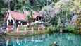 A traditional Thai building with a red roof, surrounded by lush greenery and a calm water body.